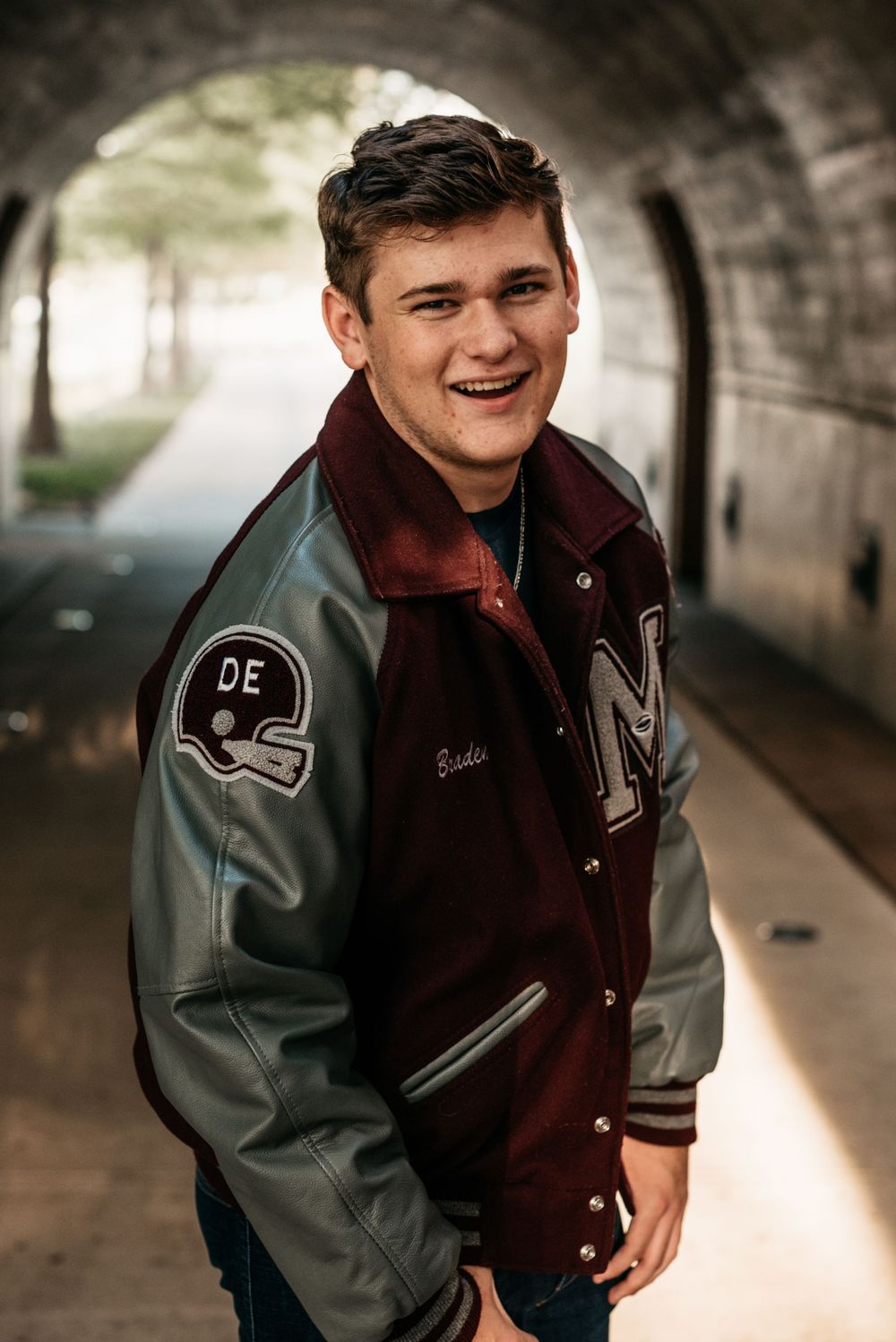 Student wearing maroon and gray varsity letterman jacket smiles while posing in tunnel with blurred background.