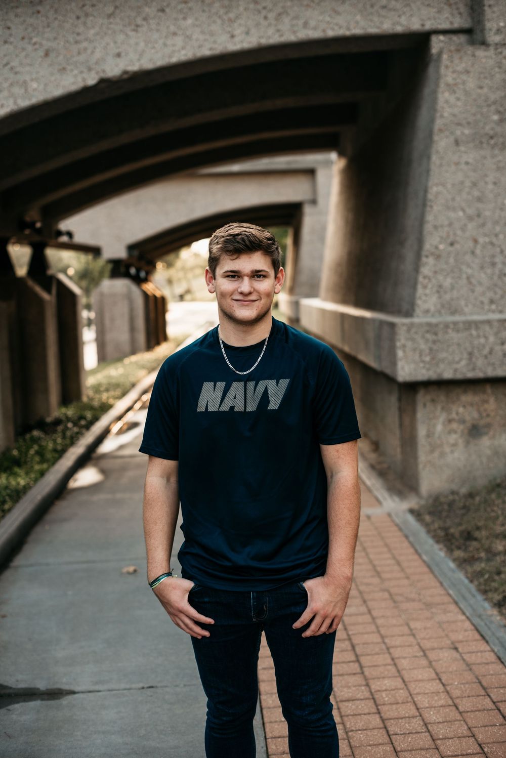 A person wearing a navy blue t-shirt with "NAVY" text stands in a covered walkway with brick pavers and arched walls.