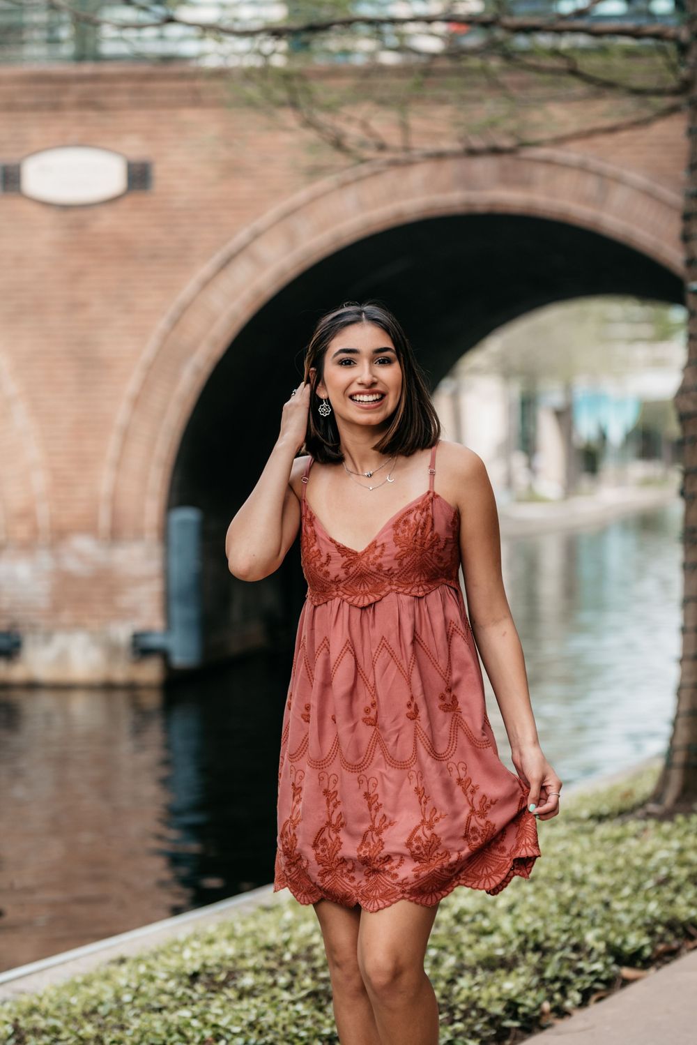 A woman in a rust-colored lace dress walks along a canal path with brick archway and water in the background.