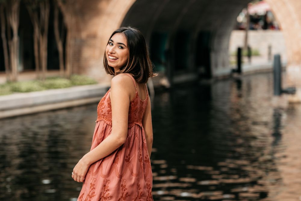 Woman in coral sundress smiles while standing by canal with stone archways in background.