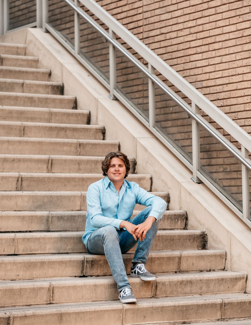 Person in light blue shirt and jeans sits casually on concrete stairs against brick wall with white handrail.
