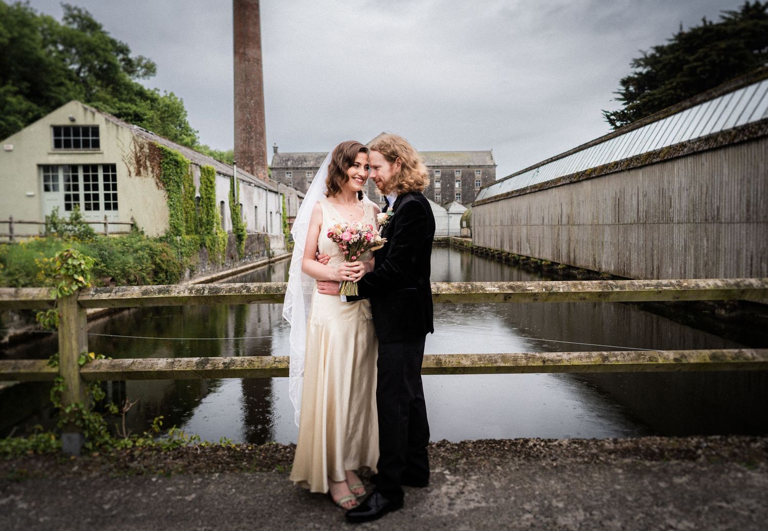 A couple embraces in front of an industrial building with a brick chimney and weathered buildings beside a waterway.