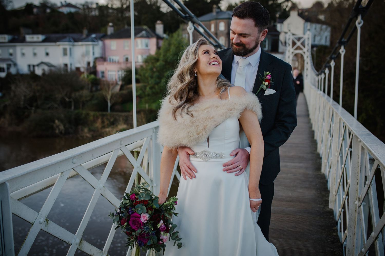Happy couple shares a romantic moment on a white bridge holding a vibrant purple bouquet at twilight.