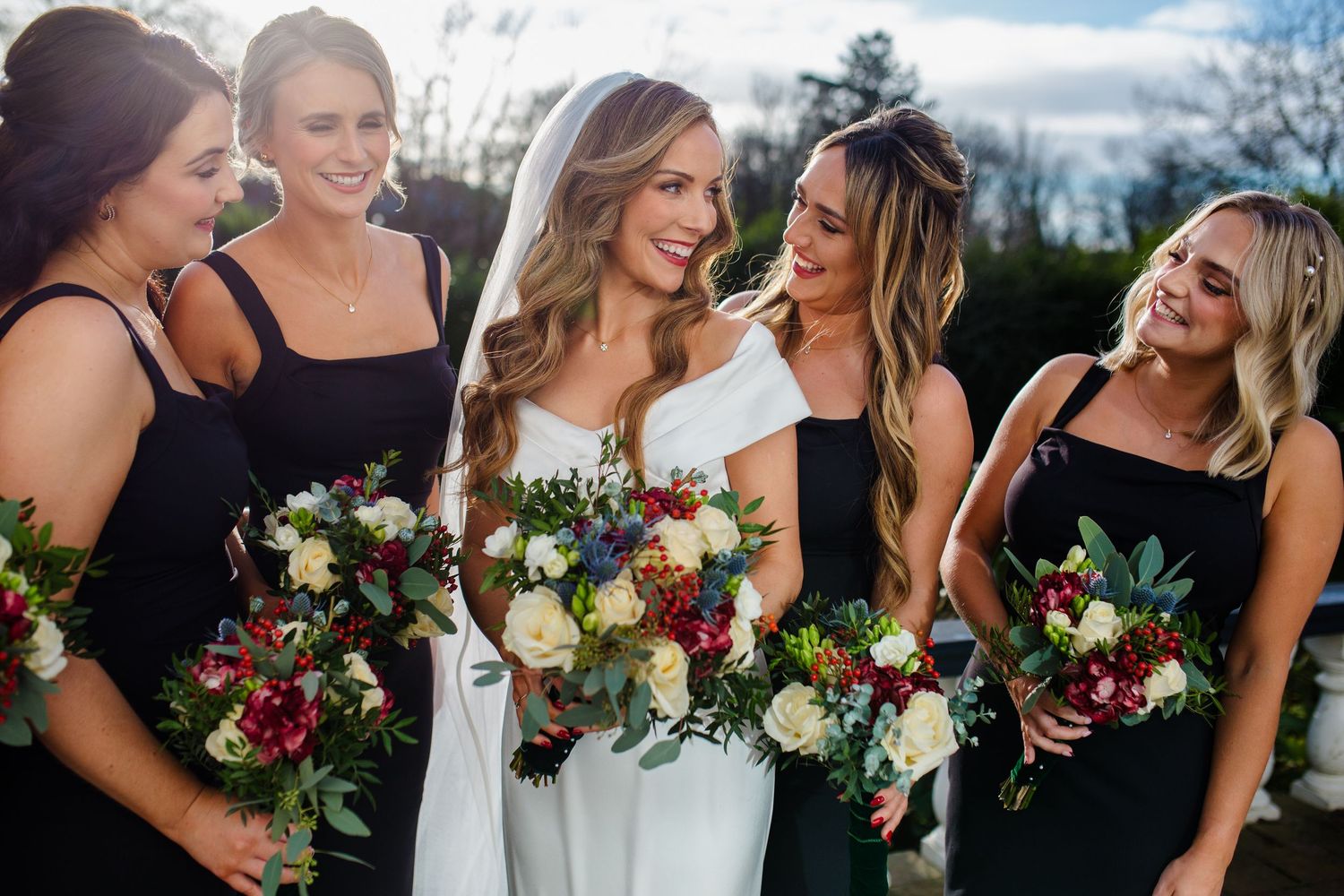 A bridal party in black dresses stands together outdoors holding colorful bouquets of burgundy and white flowers.