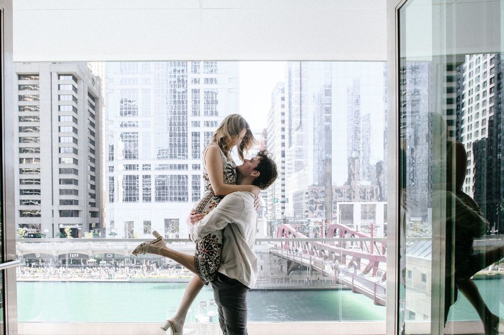A romantic couple embraces by a high-rise window overlooking a city skyline with blue water below.
