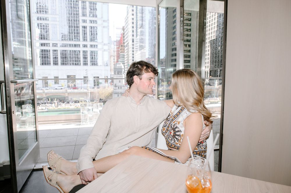 Two people enjoying drinks while relaxing in modern high-rise apartment with city views through large windows.