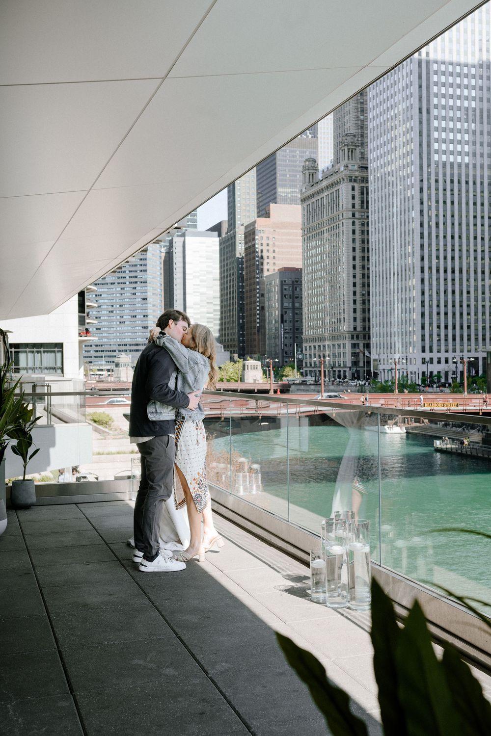 A couple embraces on a modern terrace overlooking the Chicago River with glass skyscrapers and cityscape in the background.