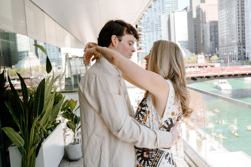A romantic couple embraces on a balcony overlooking a city river with modern buildings in the background.
