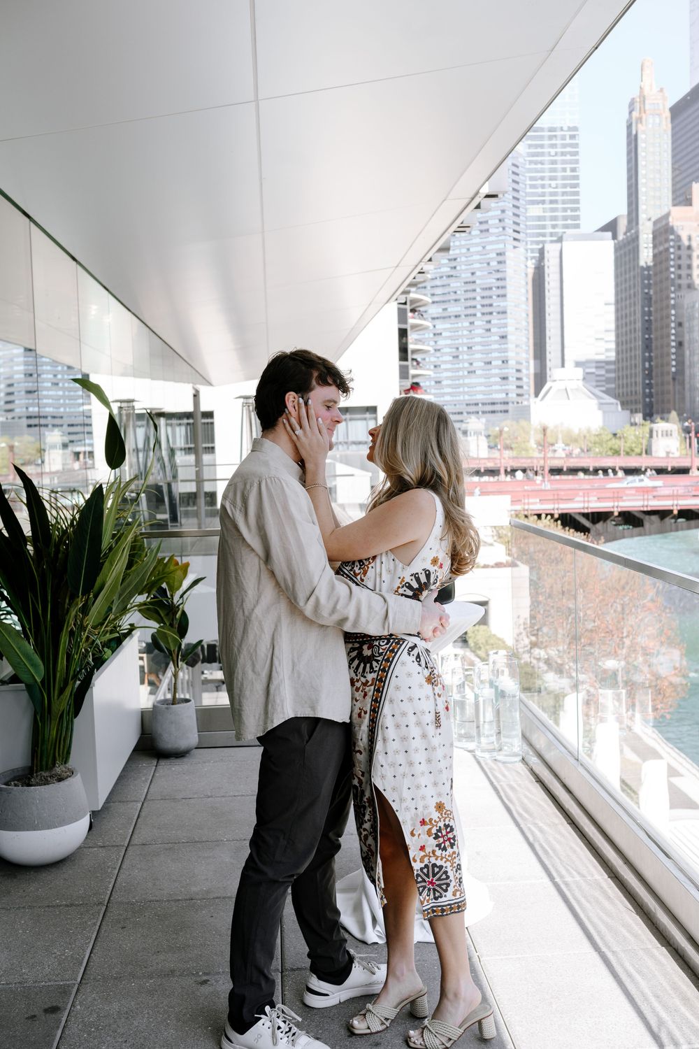 A romantic couple embraces on a modern high-rise balcony overlooking an urban cityscape with skyscrapers in the background.