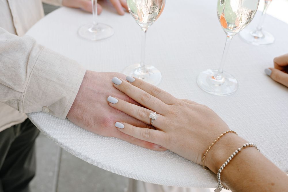 Close-up of hands displaying an engagement ring with champagne glasses on a white tablecloth.
