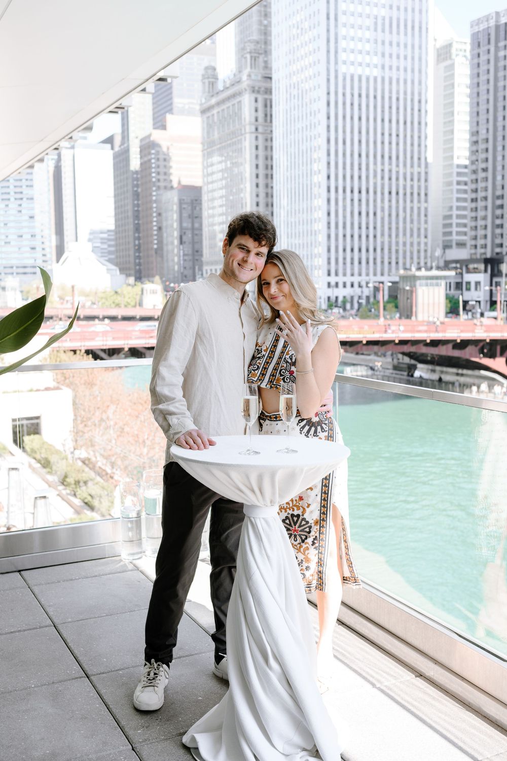 A couple poses together on a terrace overlooking the Chicago River with downtown skyscrapers in the background.