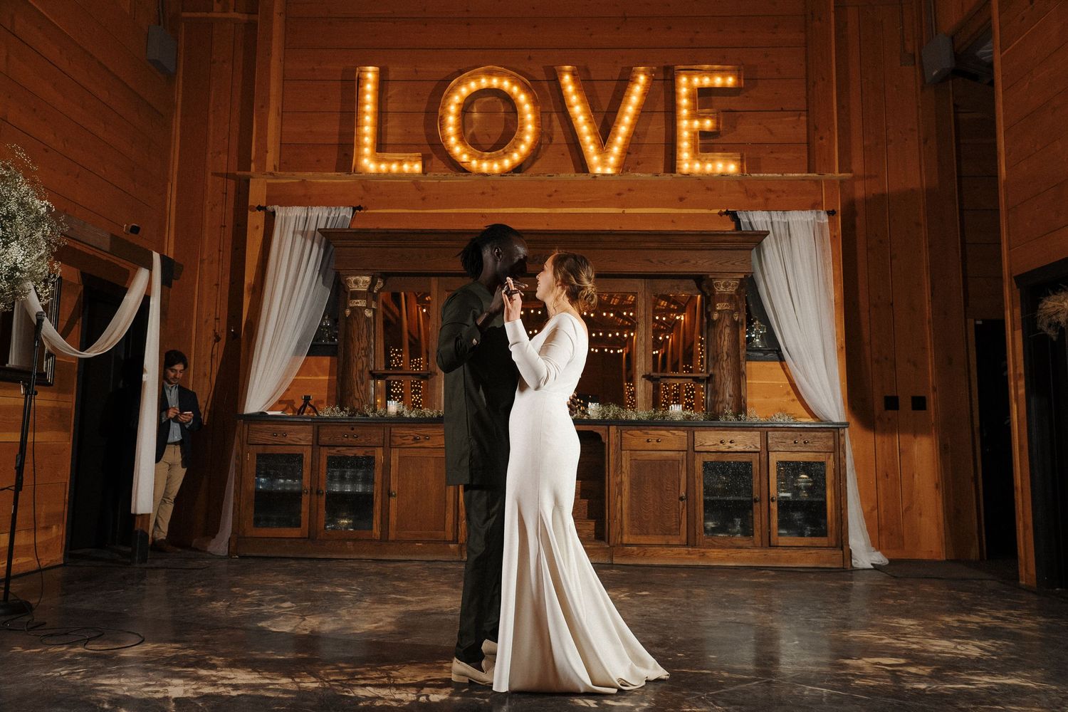 Couple dances beneath illuminated LOVE sign at rustic wooden barn venue with draped curtains and wooden bar.