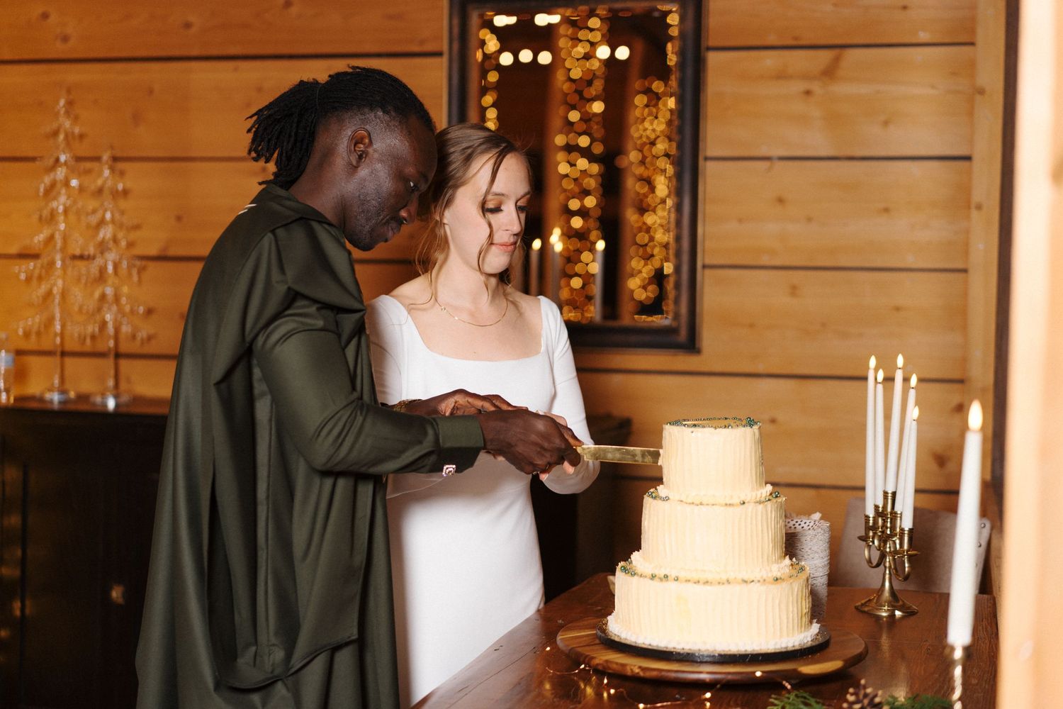 A couple cuts into their elegant three-tiered wedding cake at a rustic wooden cabin reception with candlelight and twinkle lights.