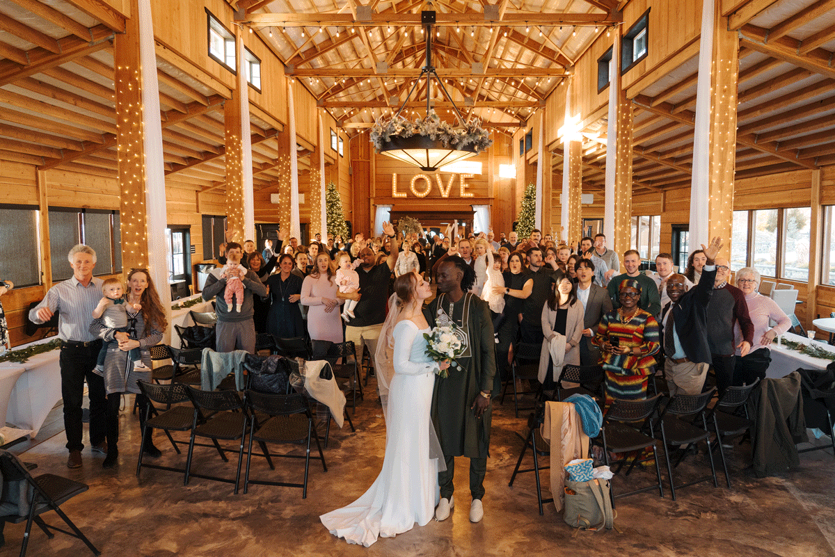 Wedding reception celebration inside rustic wooden barn venue with glowing LOVE sign and standing guests.