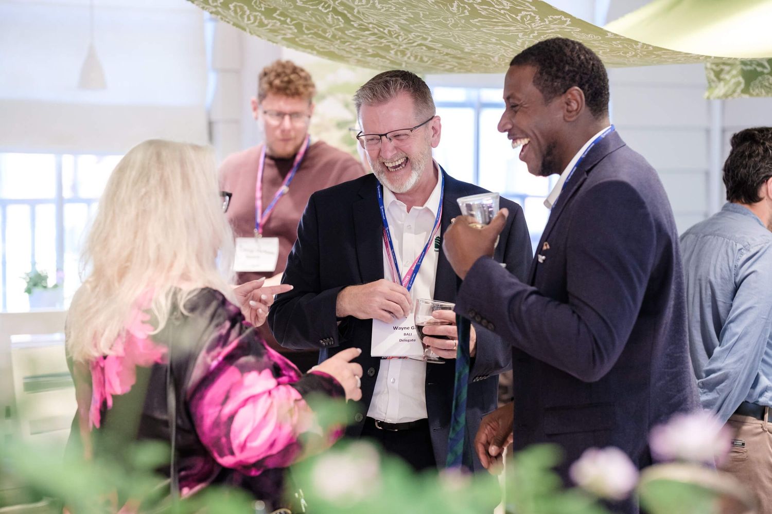 A group of professionals laughing together during a networking event, with beverages in hand at an indoor venue.