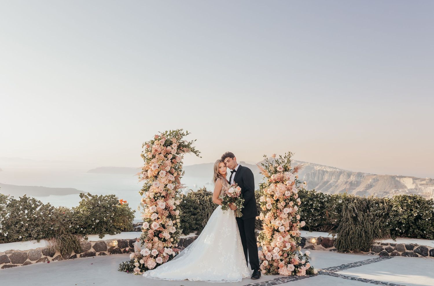 A romantic sunset wedding ceremony on a cliffside overlooking the ocean with pink floral arch decorations.