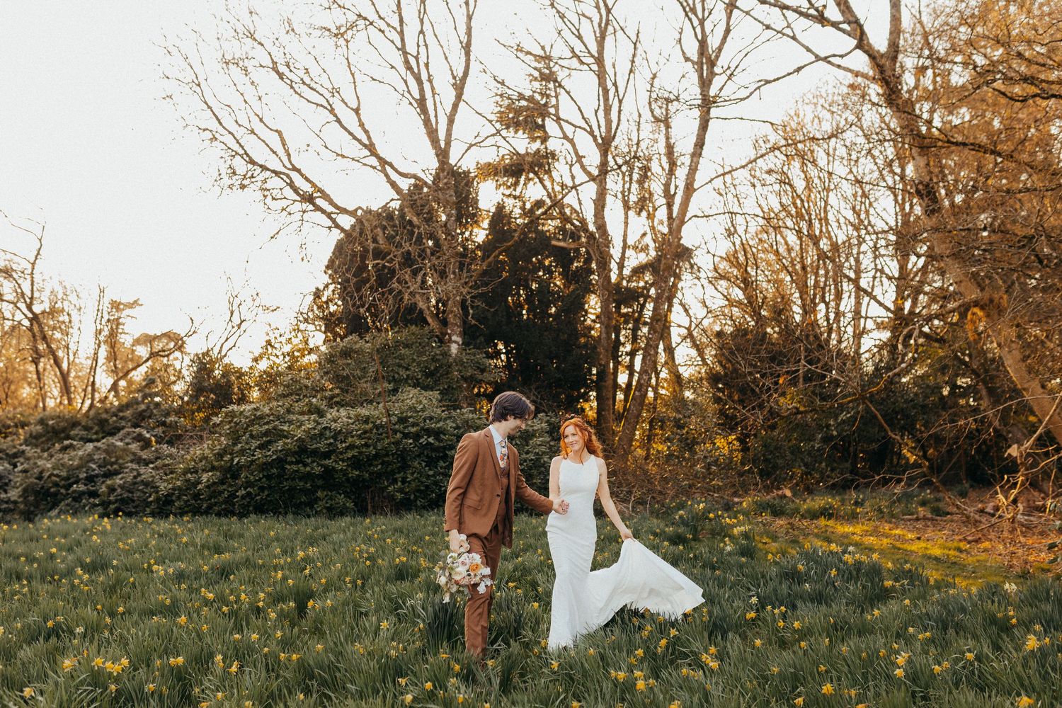 A couple in wedding attire holds hands while standing in a golden sunlit meadow surrounded by autumn trees.