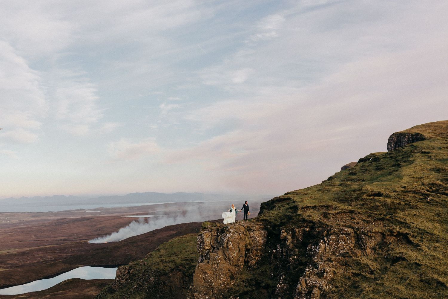 Two hikers stand atop a dramatic cliff edge overlooking a misty highland landscape during golden hour.