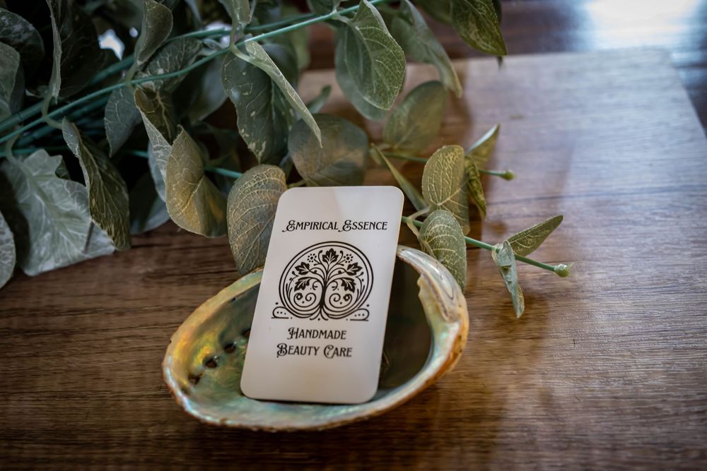 Botanical brown tea bag with sage leaves and tree logo rests on ceramic plate near green foliage on wooden table.