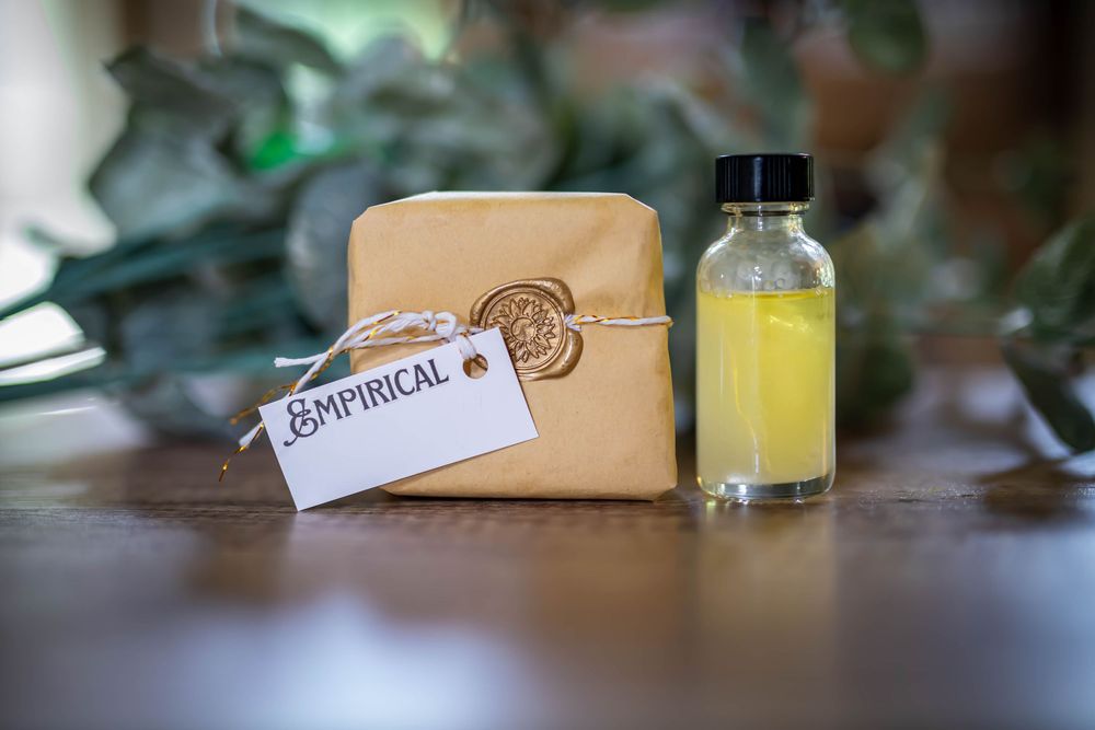 A tan leather toiletry bag with botanical tag sits beside a glass bottle of yellow liquid on wooden surface.