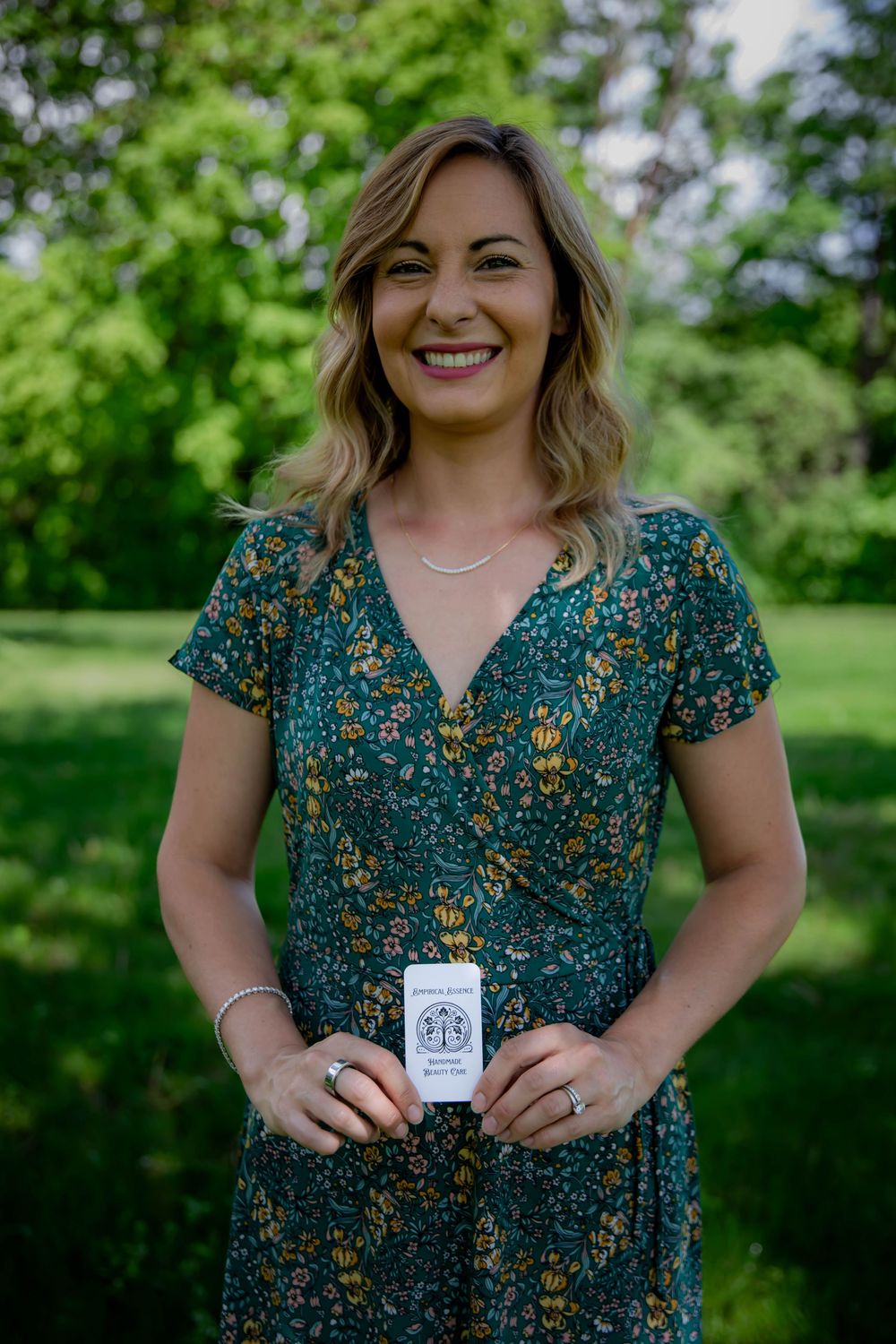 A person holding a small product container stands outdoors wearing a floral patterned teal dress against a green natural background.