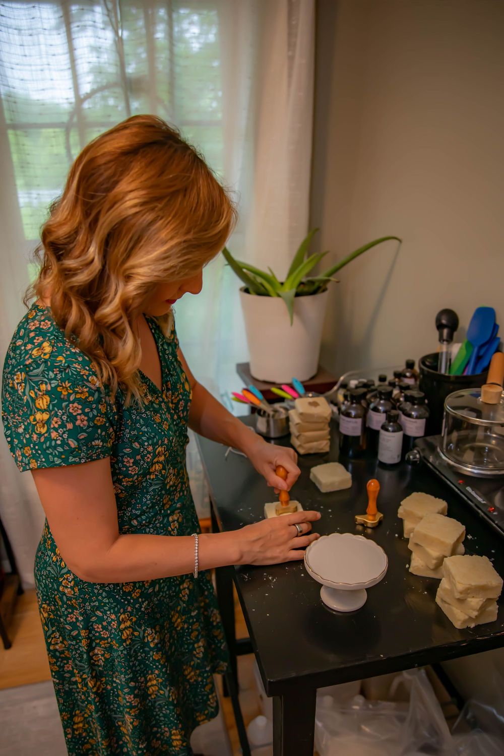 A person in a floral green dress works at a table with various bottles and jars arranged on a black surface.