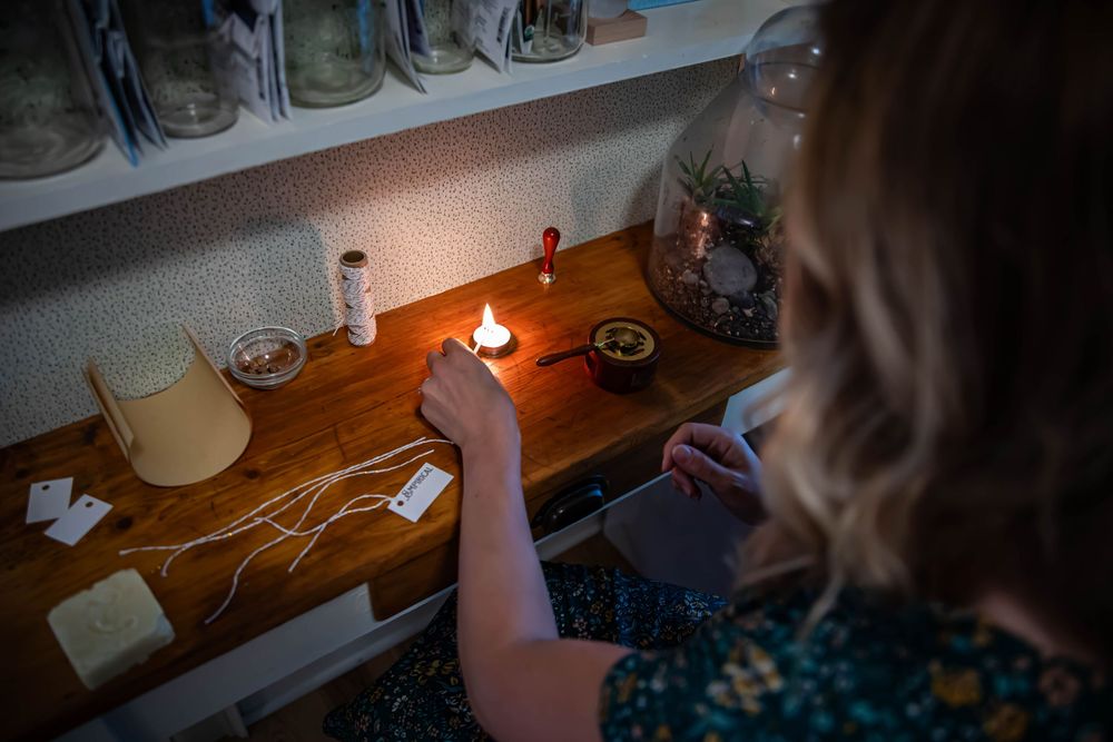 A dark scene showing hands lighting a candle on a wooden shelf with glass jars.