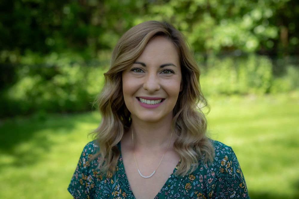 A woman in a teal floral dress poses outdoors against a green natural background.