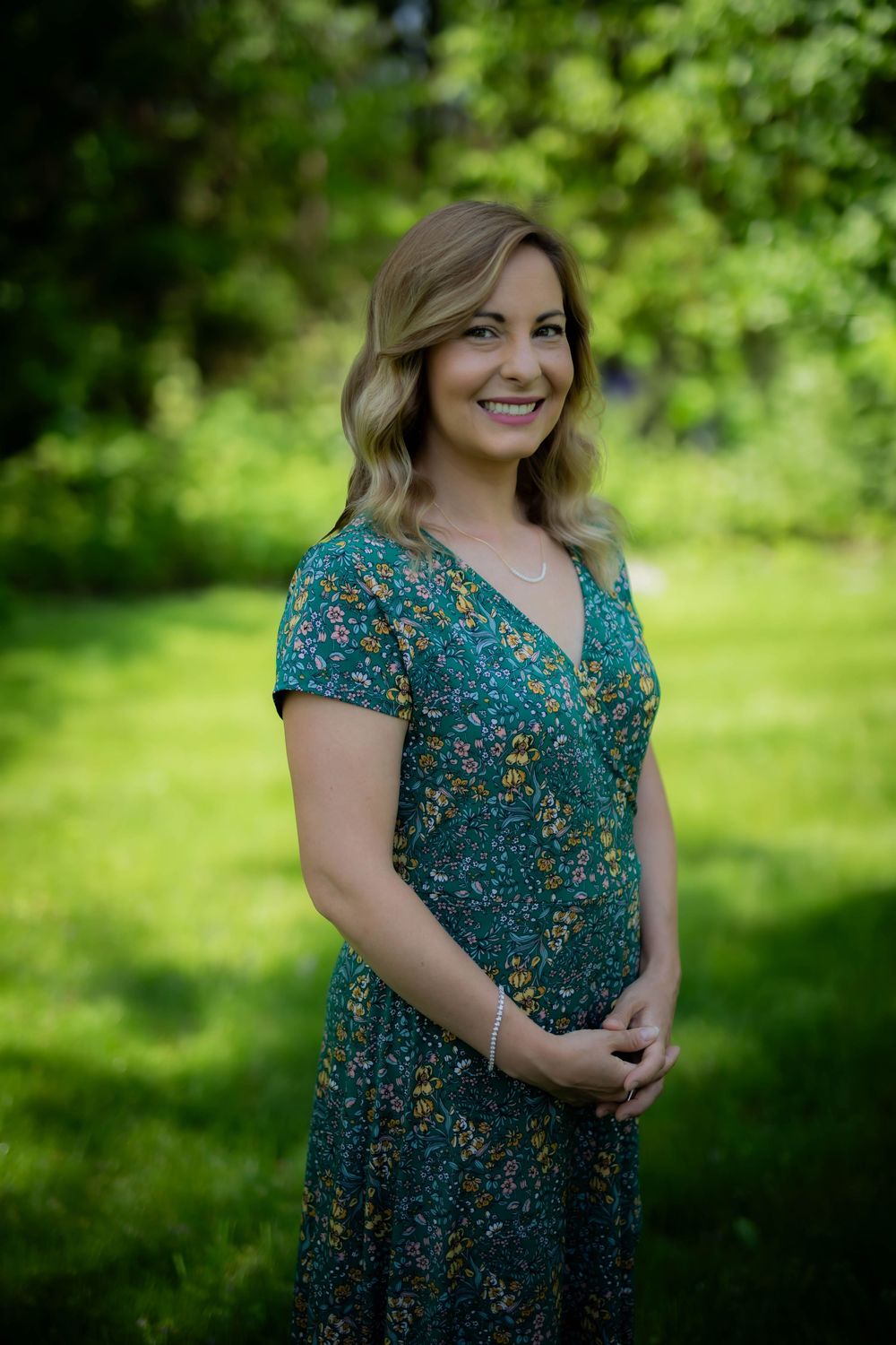 A woman in a floral teal dress stands smiling in a lush green garden setting.