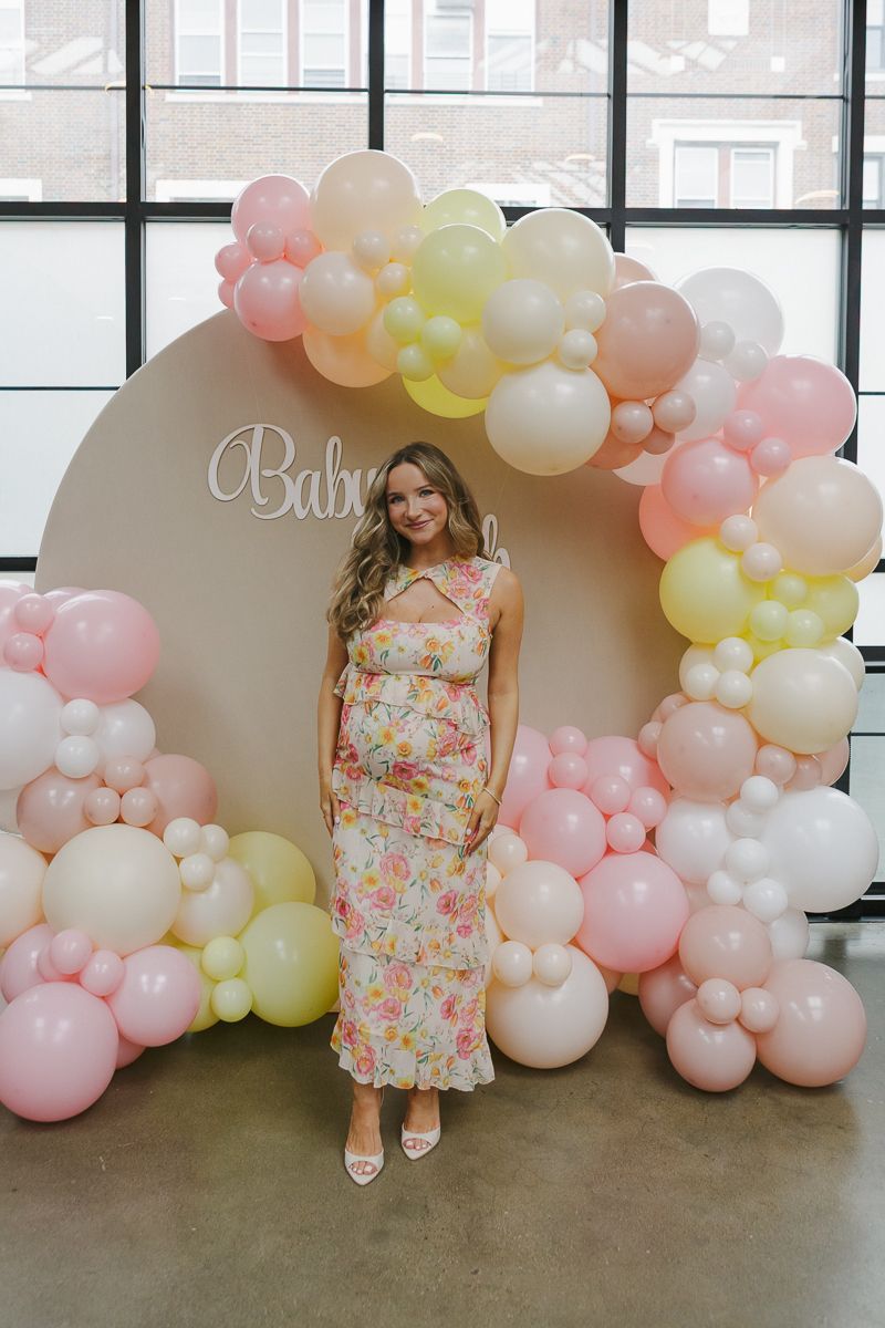 Radiant mom-to-be in a floral dress posing in front of stunning pink balloon decor, captured by NYC baby shower photographer Paula Brearley.