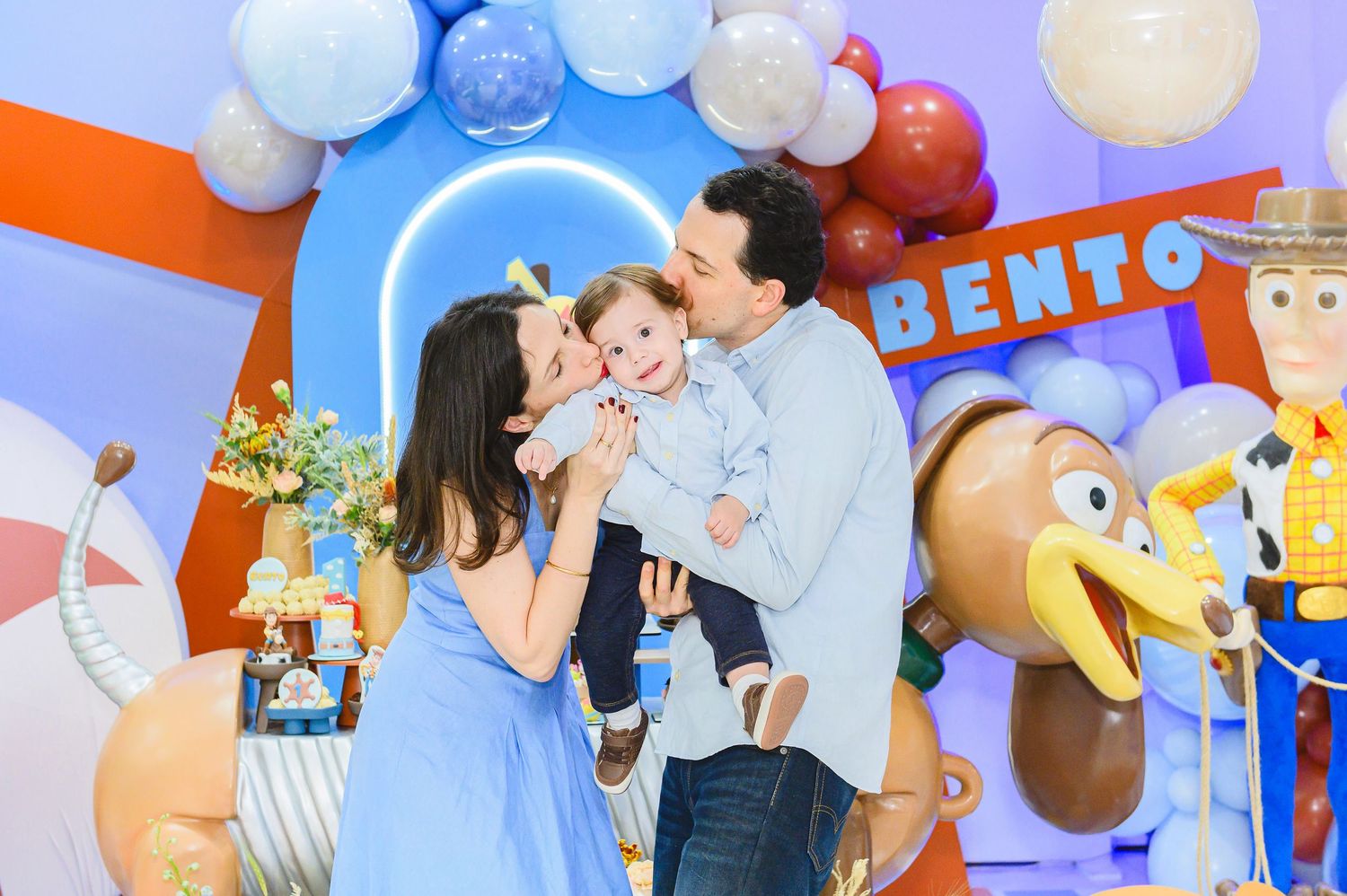 A family celebrates at a colorful Toy Story themed birthday party with Woody decorations and blue and white balloons.