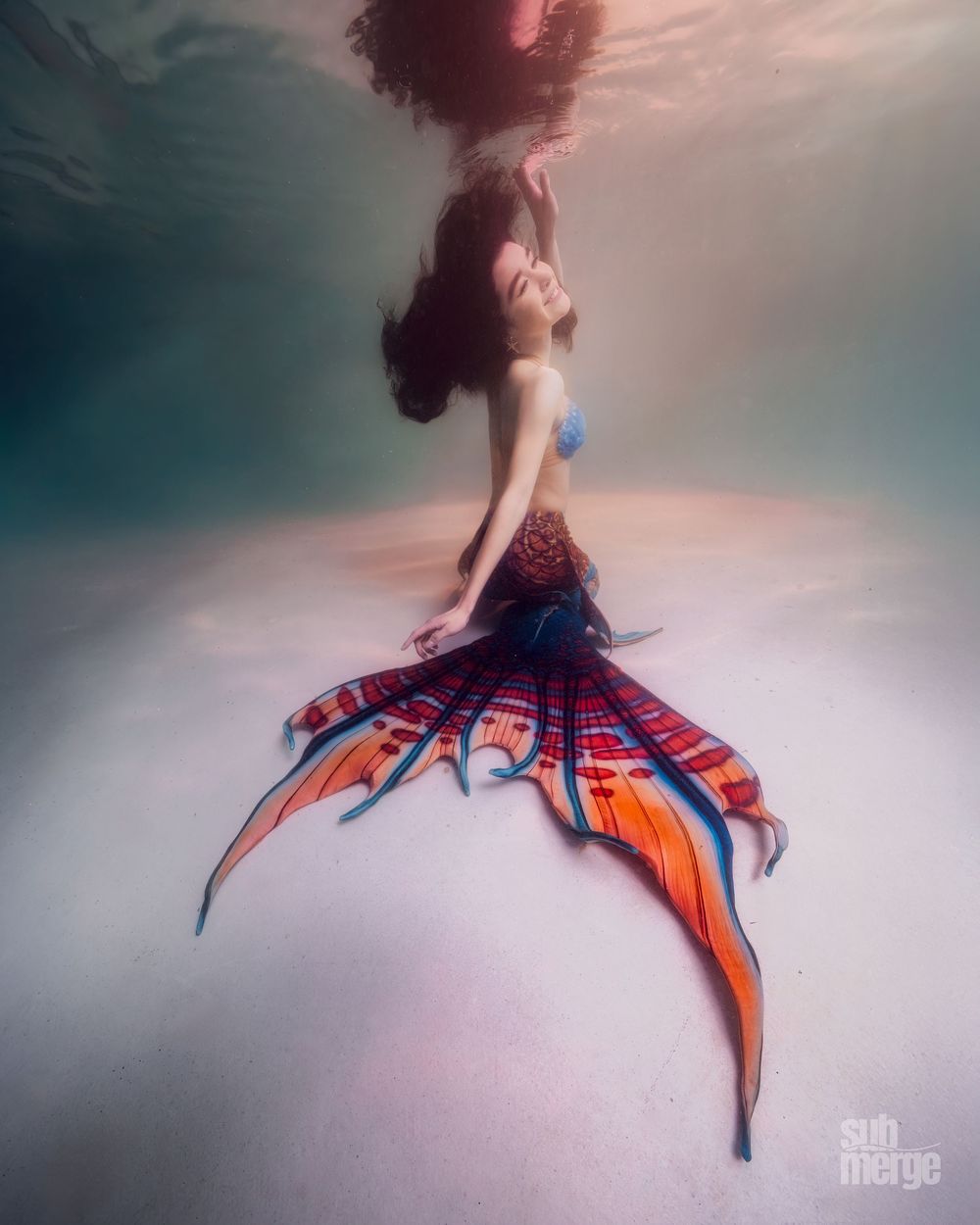 Ethereal underwater photo showing a figure in a flowing orange and red dress floating gracefully in dreamy aqua water.