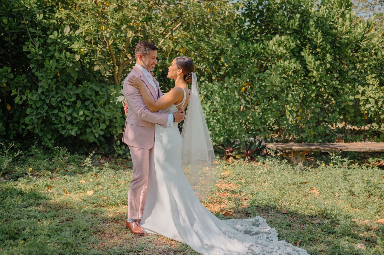 A couple embraces outdoors during their wedding ceremony, with the bride wearing a flowing white gown against green foliage.