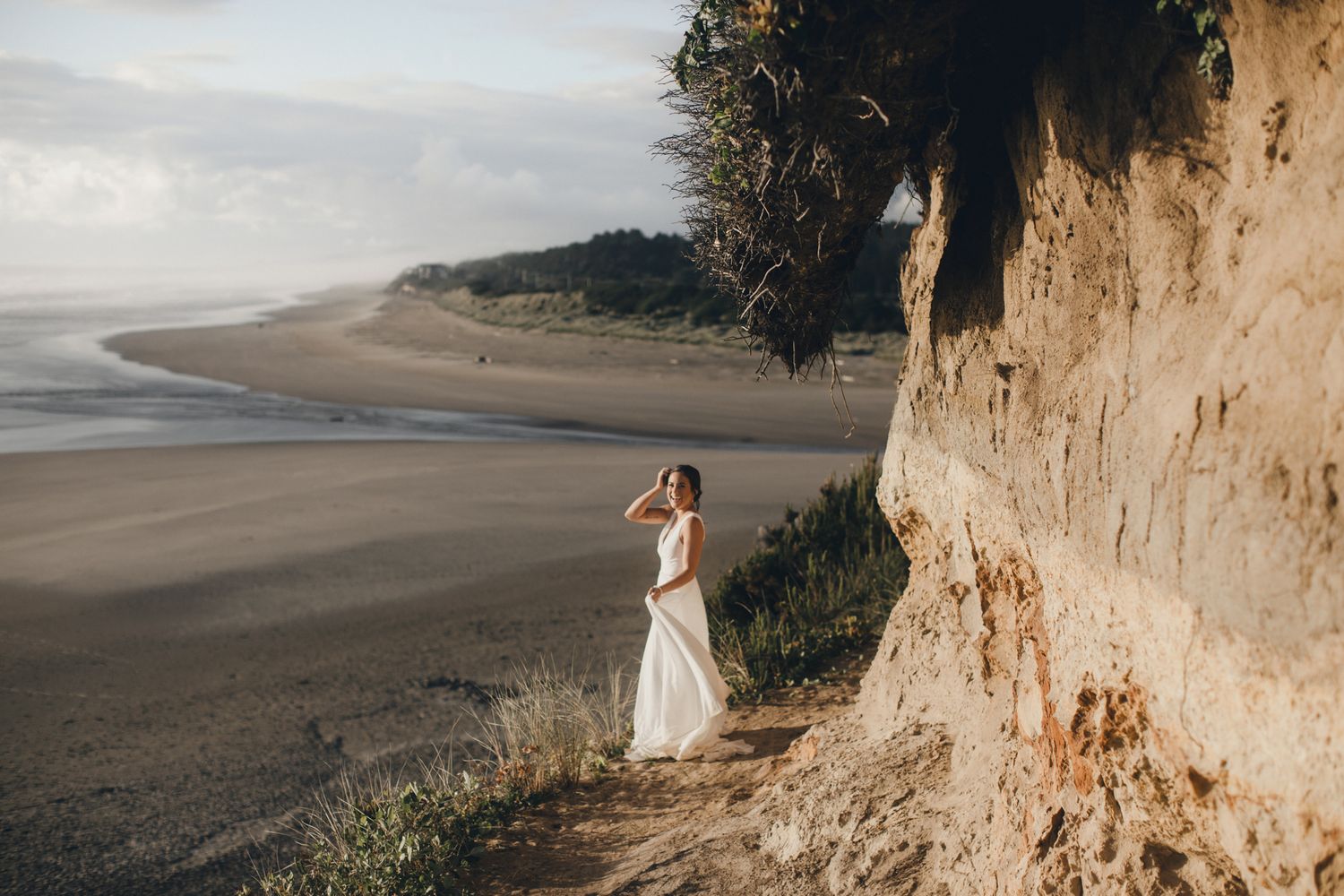 Someone in a white wedding dress stands on a beach near dramatic coastal cliffs at sunset.