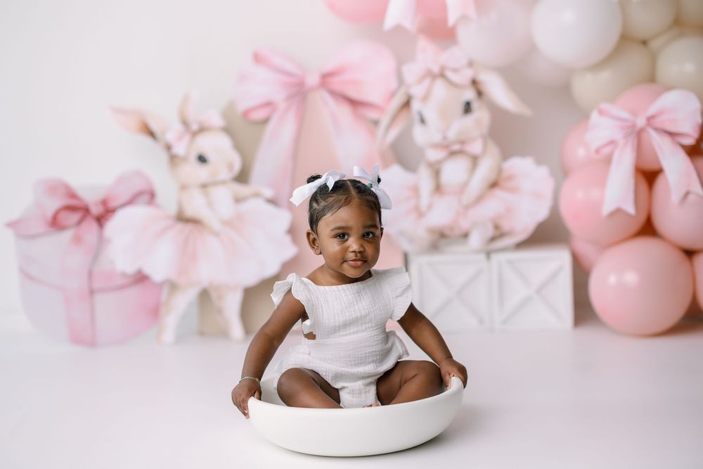 Baby sits in white bowl against pink backdrop with balloons and gift boxes for first birthday photoshoot.