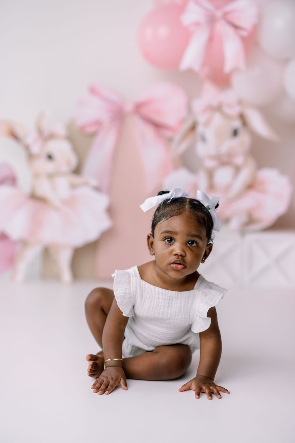 Sweet baby portrait with pink balloon decorations and bunny plush toys against pastel studio background.