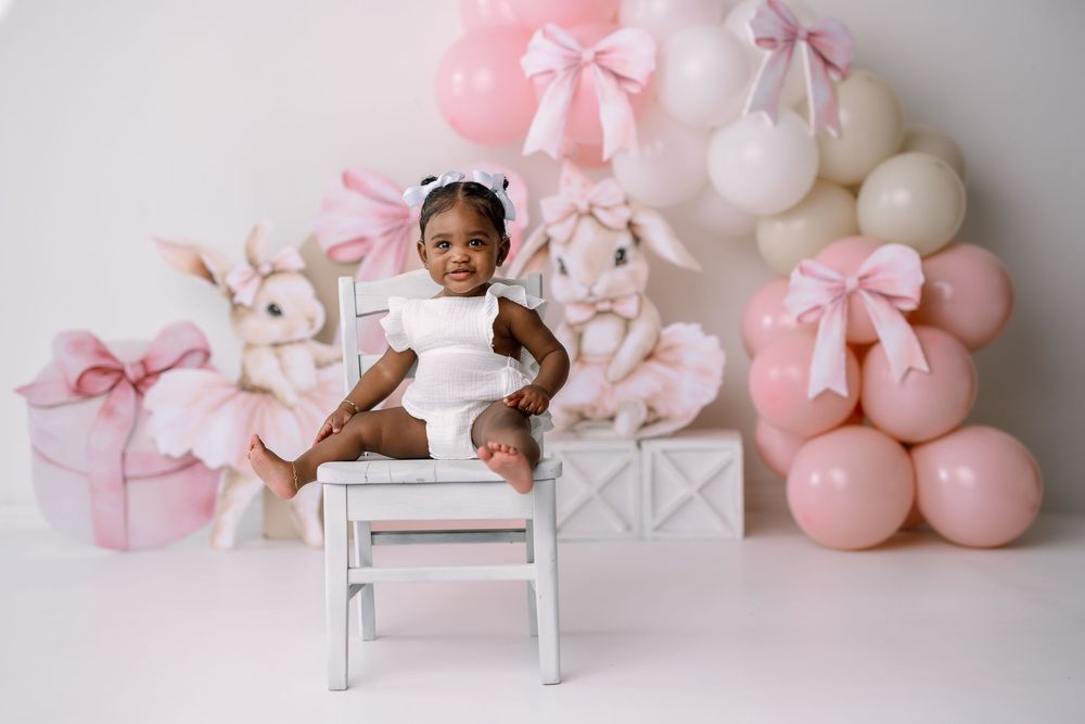 Festive pink and white party setup with balloons, presents, stuffed bunnies and white chair against neutral backdrop.