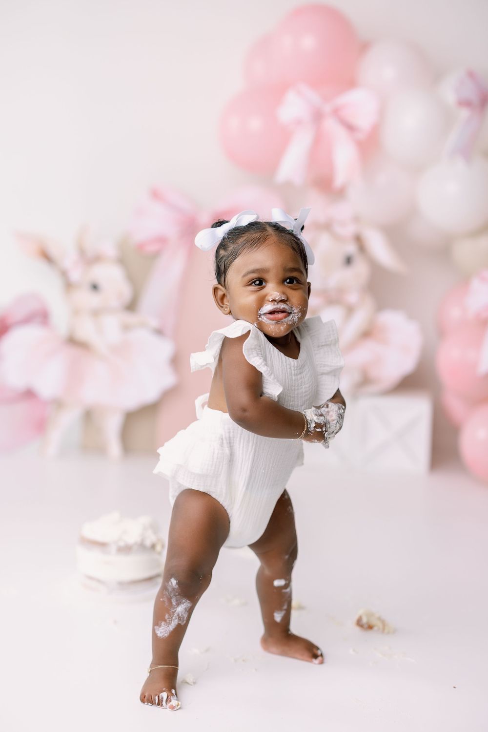 Baby in white dress crawls and plays among pink and white balloons during a delicate spring photoshoot.