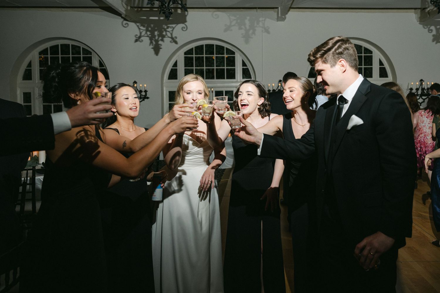 A wedding party group shares a celebratory toast together in a formal venue with arched windows.