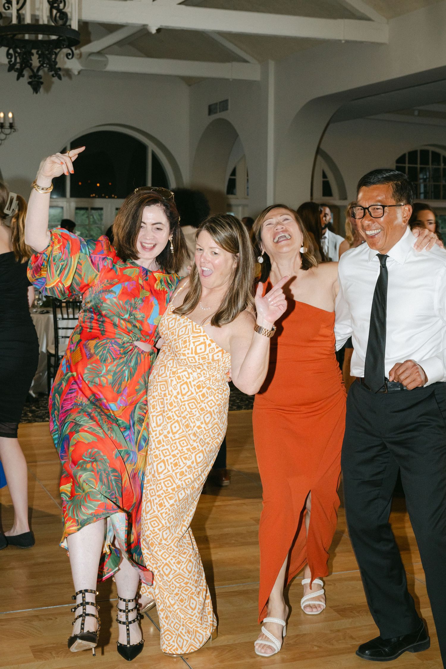 Party guests dancing together on a wooden dance floor at an indoor event venue with arched architecture and chandeliers.