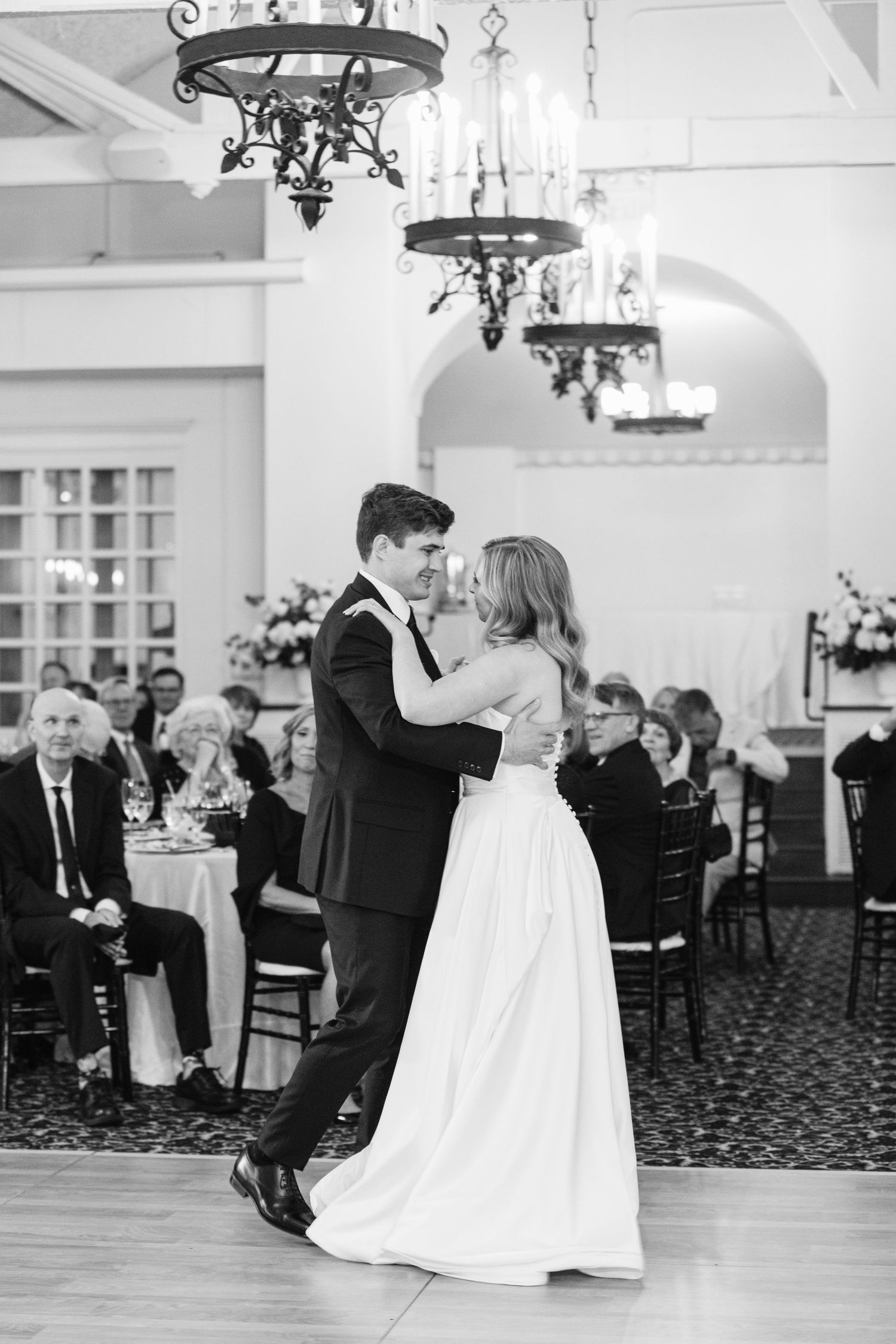 A couple shares their first dance at a traditional wedding reception under elegant chandeliers in a grand ballroom.