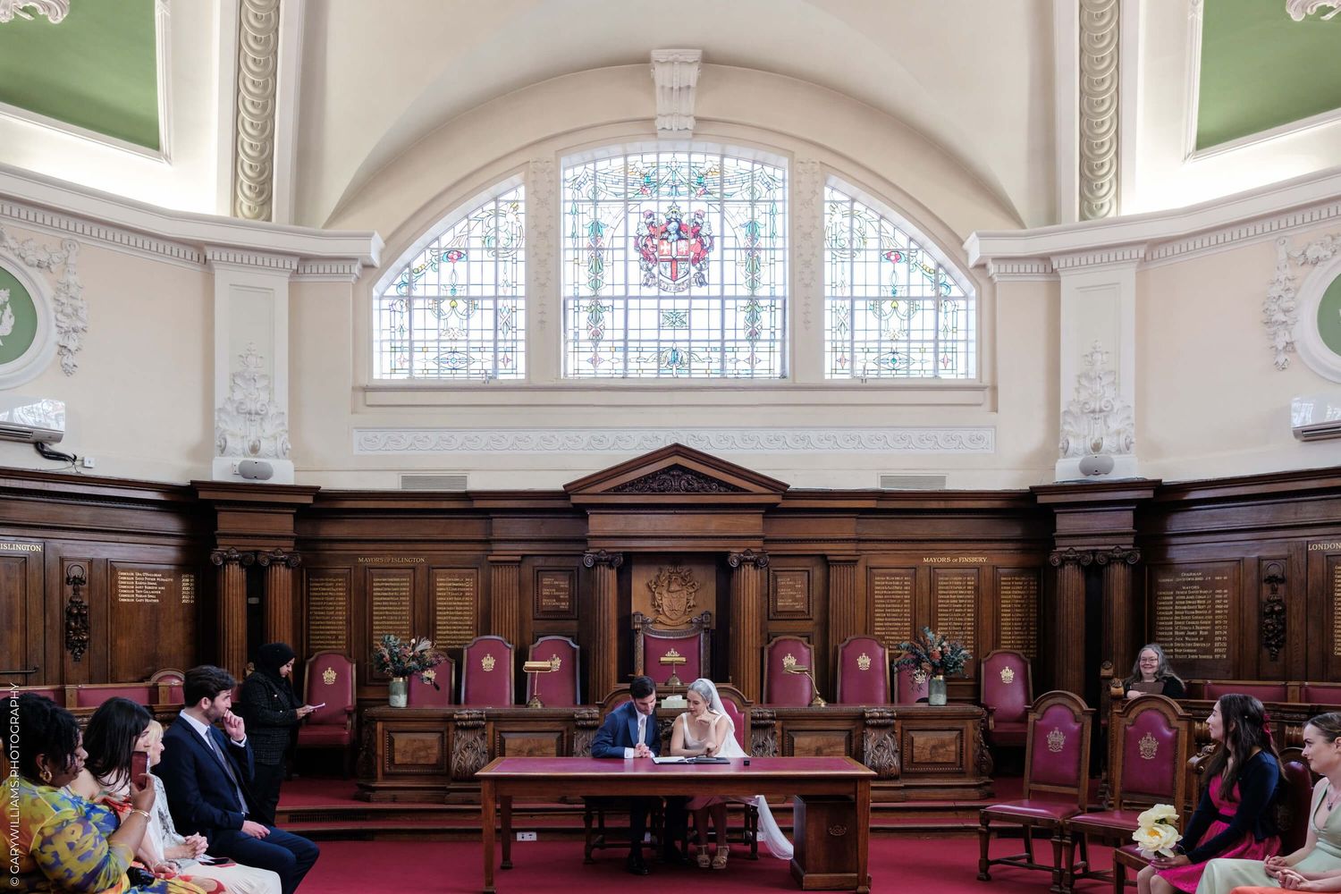Meeting or ceremony taking place in historic wood-paneled council chamber with arched stained glass window and ornate architecture.