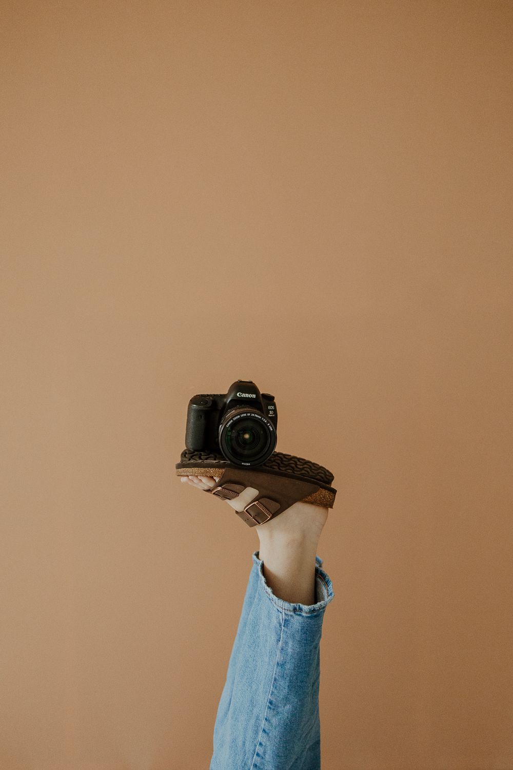 Foot in denim holds up a black DSLR camera against beige background in minimal photography composition.