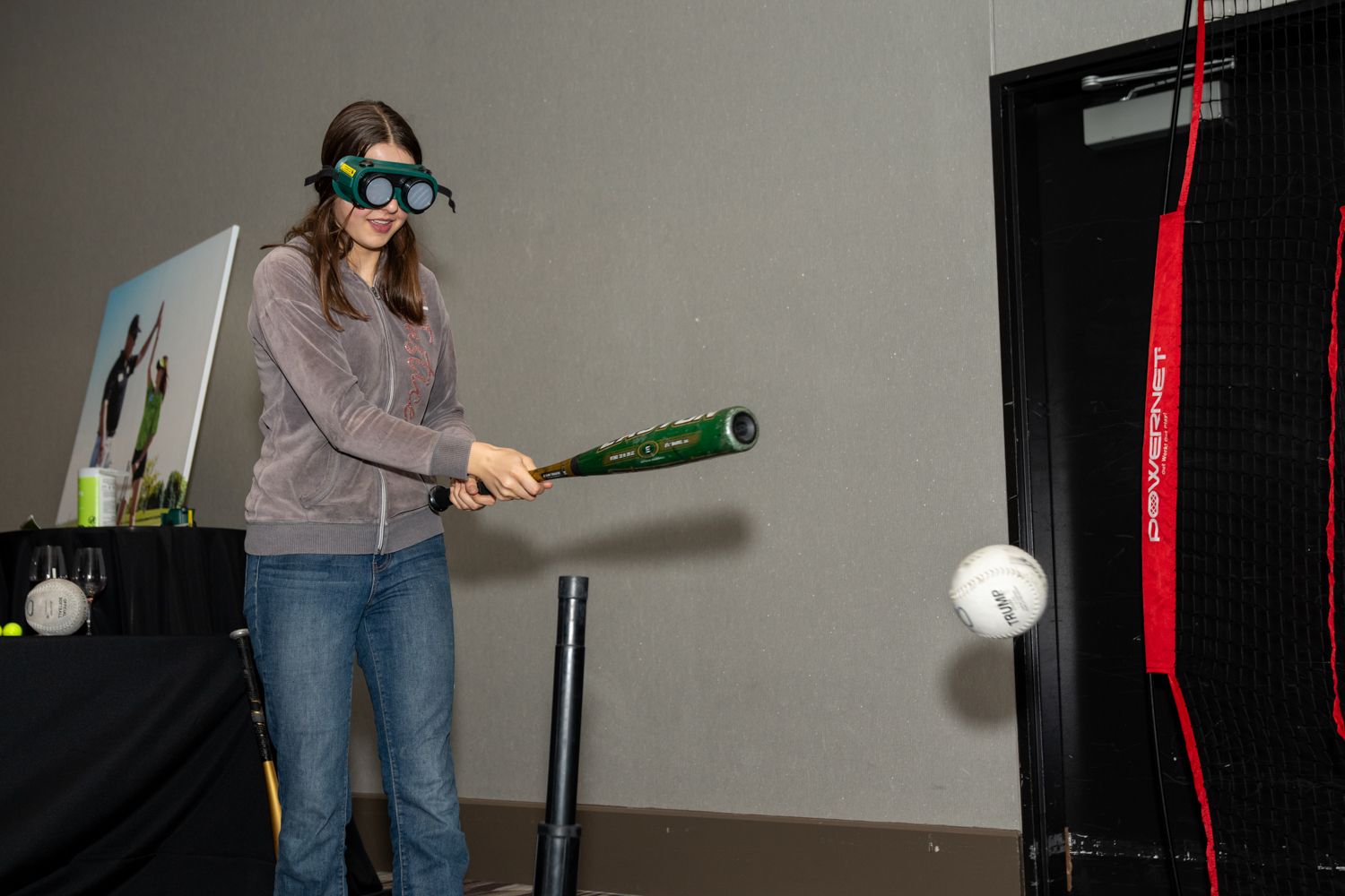 Person wearing goggles swings virtual bat at suspended ball in gaming setup.