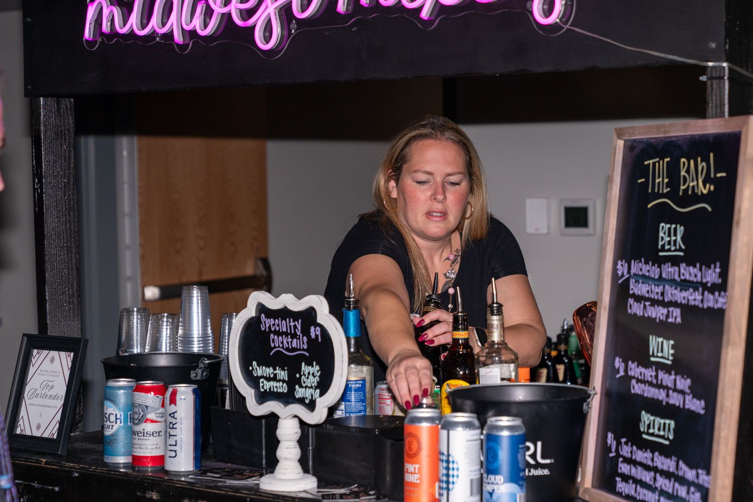 Bartender behind counter serving drinks at nighttime event with neon signage.