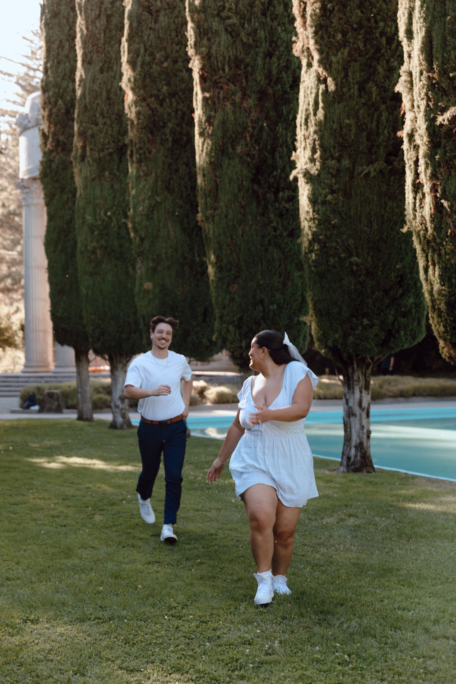 Two people in white outfits jog together along a path near cypress trees and a pool on a sunny day.