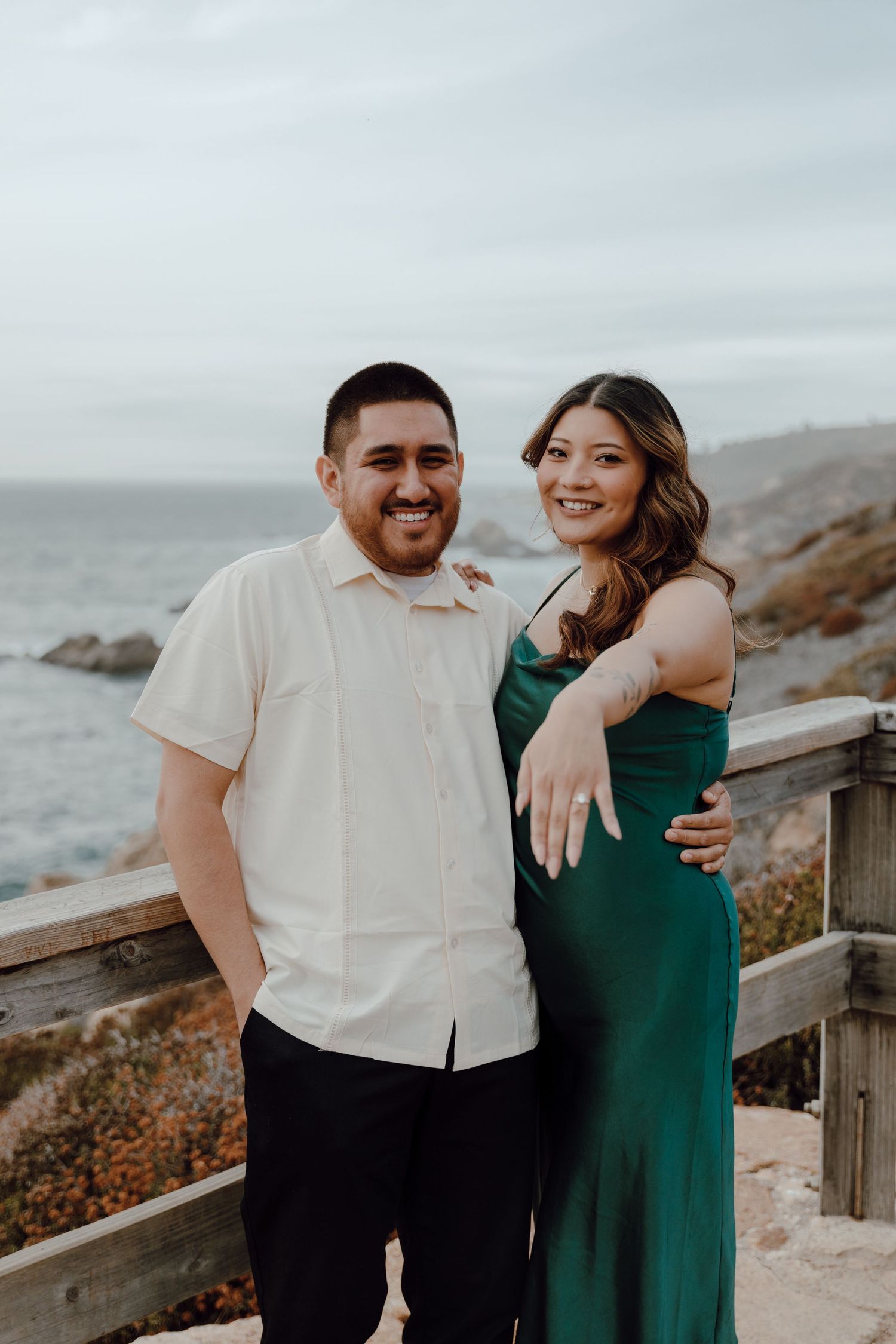 A couple poses together on a coastal overlook, with the woman wearing an elegant emerald green dress.