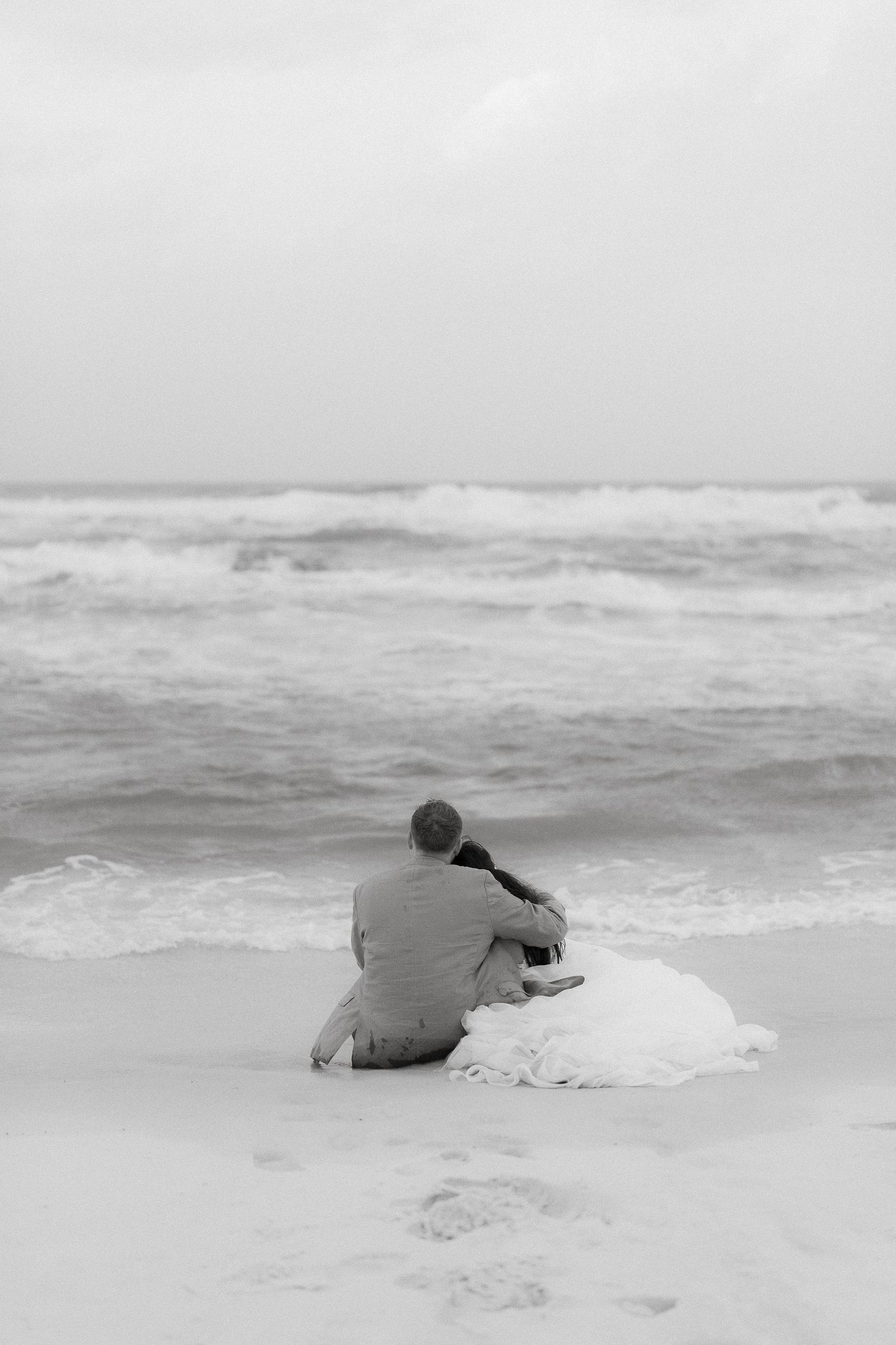A solitary person in casual attire sits on the sandy beach near crashing waves along a misty shoreline.