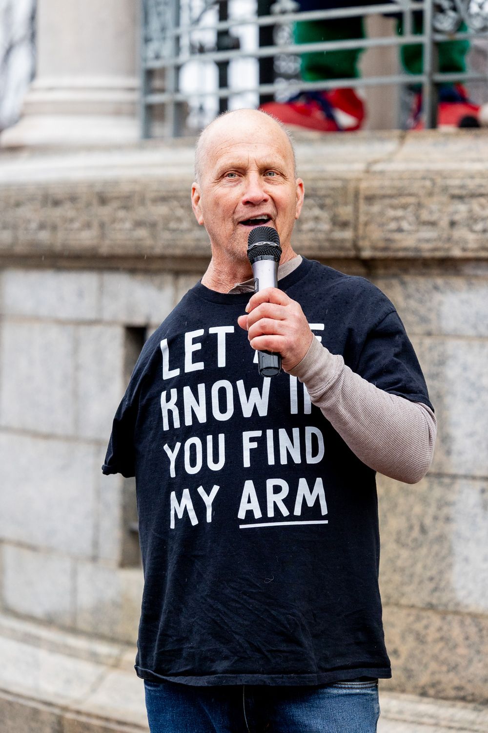 A person with a microphone wearing a black t-shirt with text that reads 'LET KNOW IF YOU FIND MY ARM' speaks outdoors.