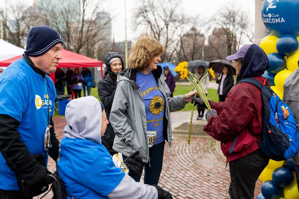 A group gathers at a community event with blue jackets, yellow balloons, and red tents on a brick pathway.